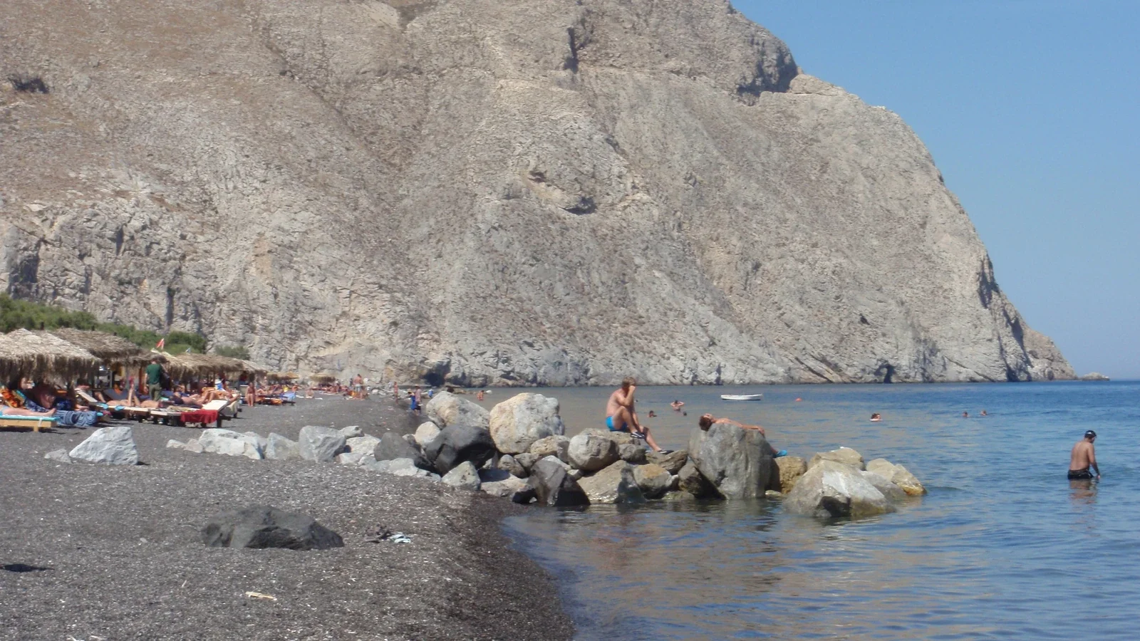 Тёмный пляж Периссы с отдыхающими и камнями у воды под высокой светлой скалой